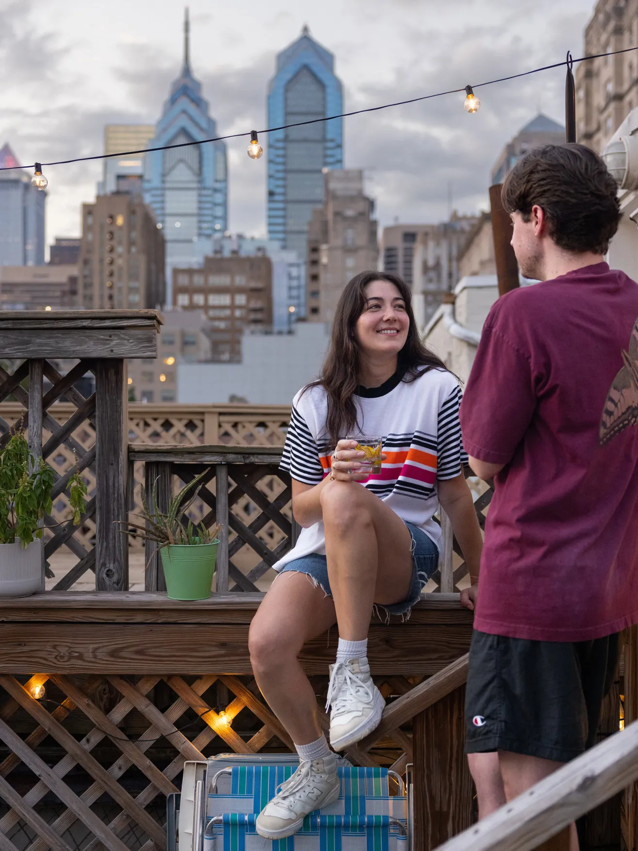 With the Philadelphia skyline behind them, Tovah Kaiser sits on a wooden railing holding a drink and smiling as she chats with her boyfriend. Hey’re both wearing T-shirts and shorts, and see, happy and relaxed. 