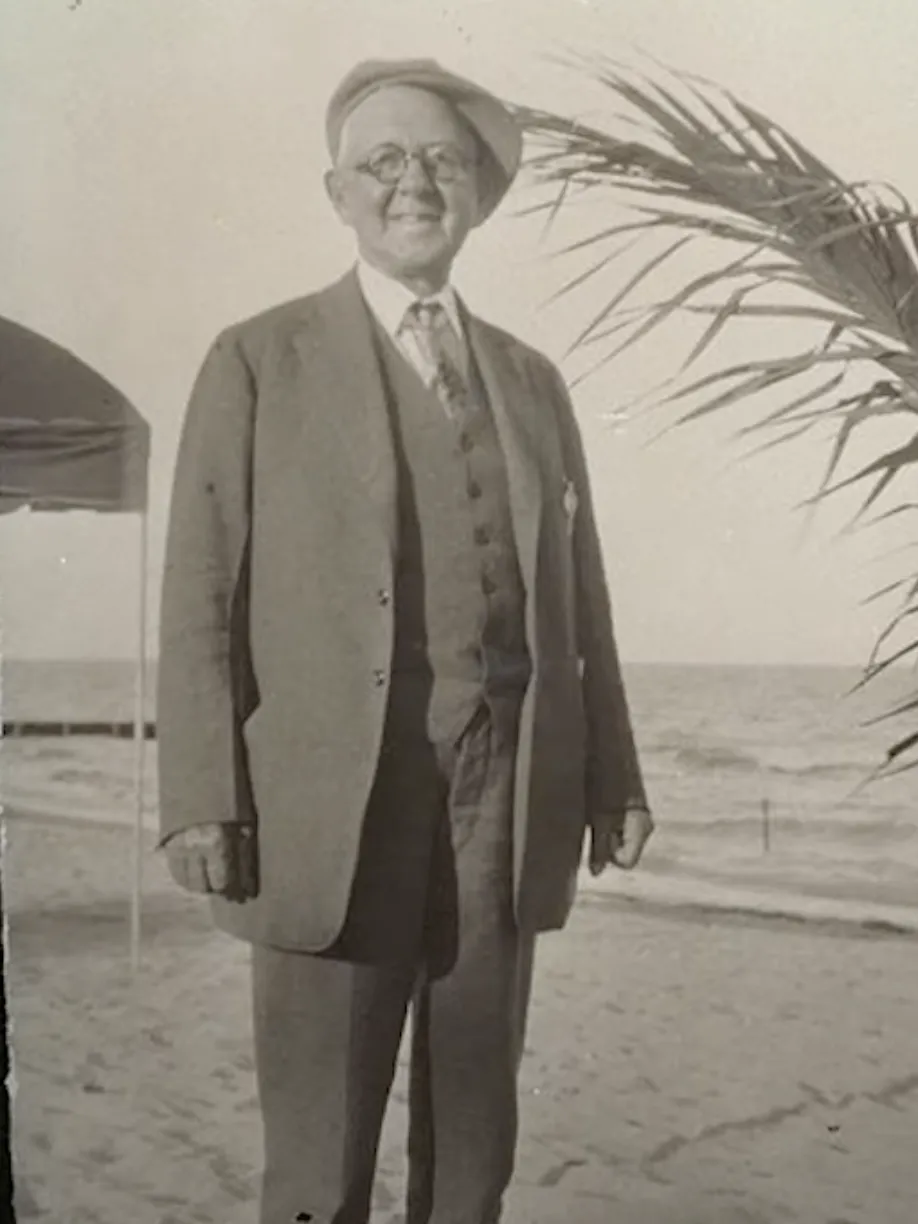 In the 1920s a man wearing glasses and a full suit stands on a beach