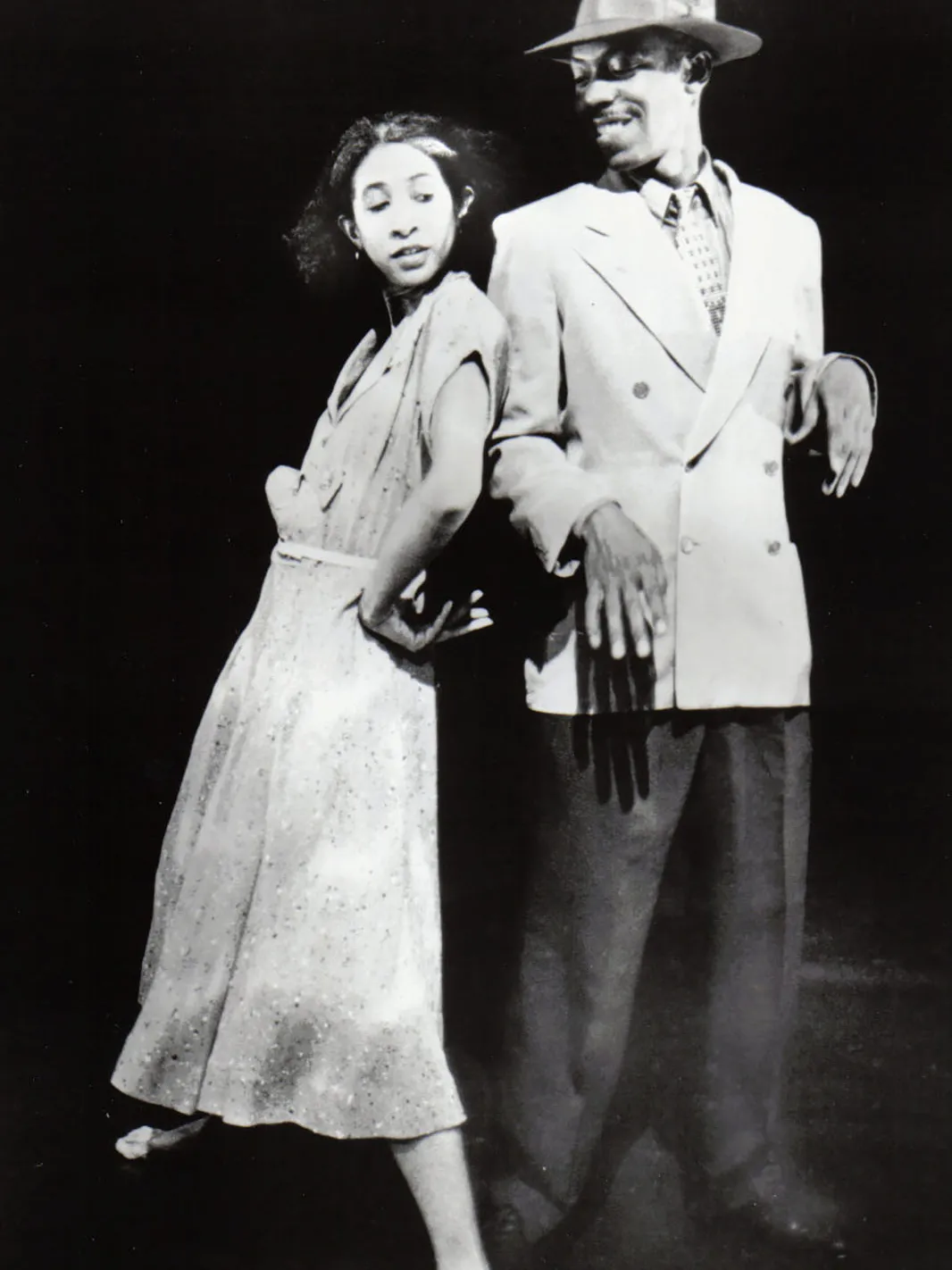 On stage, a younger Dianne playfully leans her back against a man's arm. He is a tall black man wearing a fedora and suit; she has on a shirt dress. He’s smiling as he looks down at her and she’s looking over her shoulder at him, but not at his face.