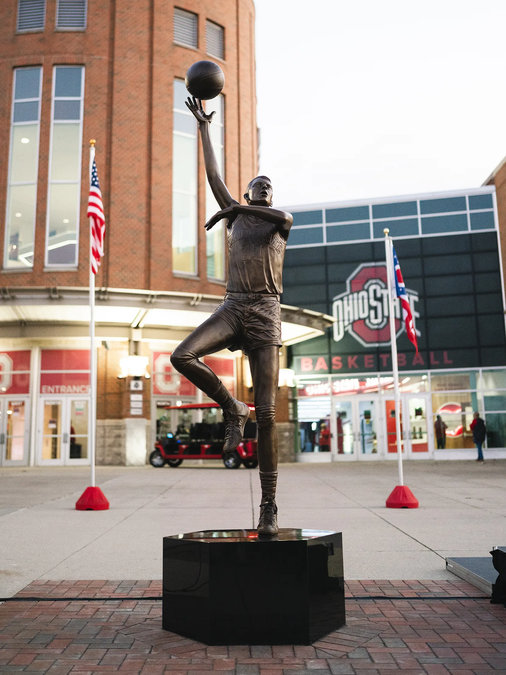 The statue of Jerry Lucas outside the Schottenstein Center is bronze and bigger than life size. Jerry lifts his right leg, extending his left with his weight there. His right arm is almost fully extended but slightly rounded as he rolls the basketball off his fingertips. His left arm is ben loosely in front of his torso. His open-mouthed expression shows his focus.