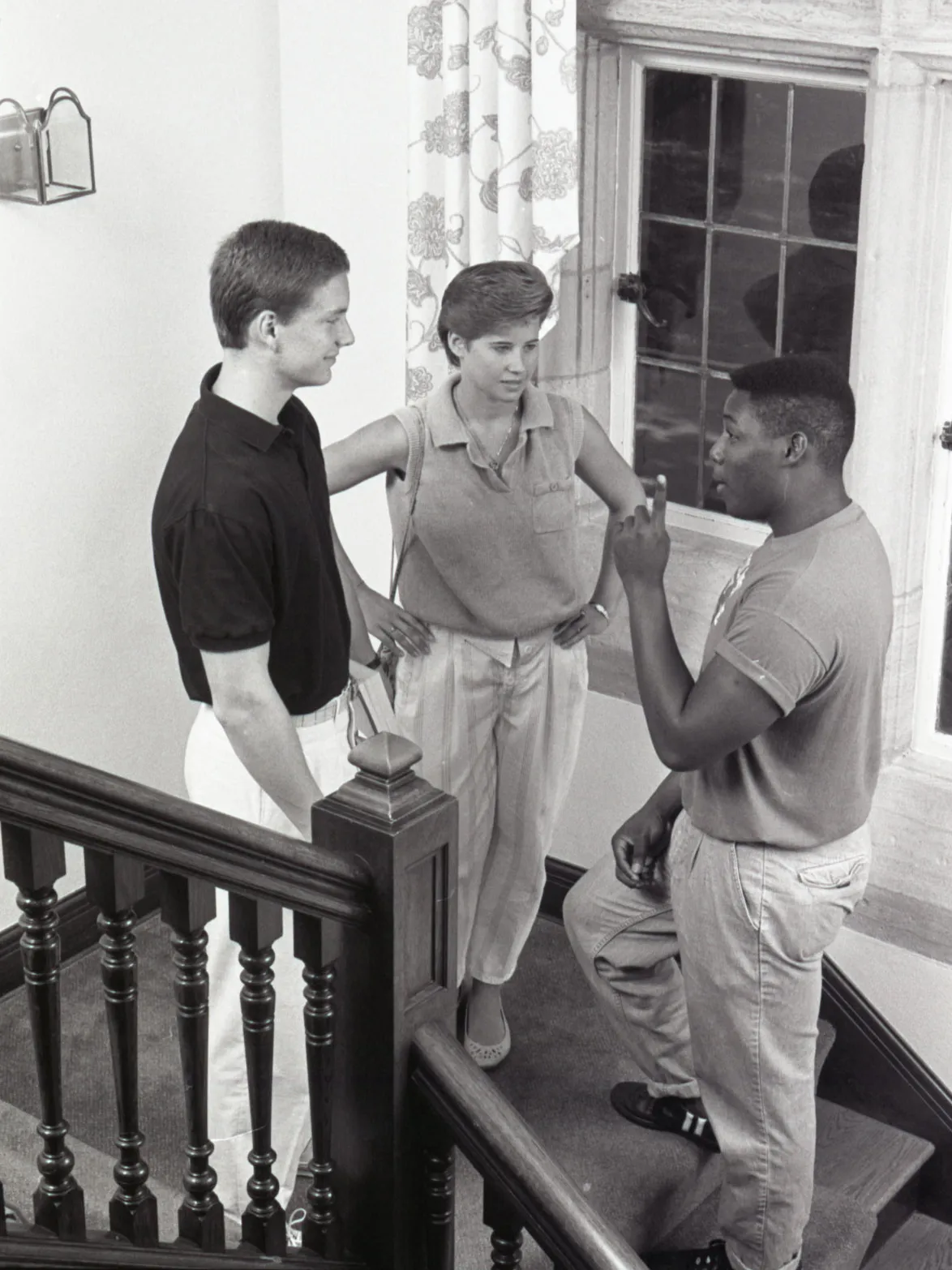On a staircase landing, three college students engage in conversation. A large window with patterned curtains is behind them, and the wooden staircase features turned balusters and a decorative newel post. Of the three, one is woman and two are men; one is Black and two are white. They’re dressed in styles common in the 1980s.