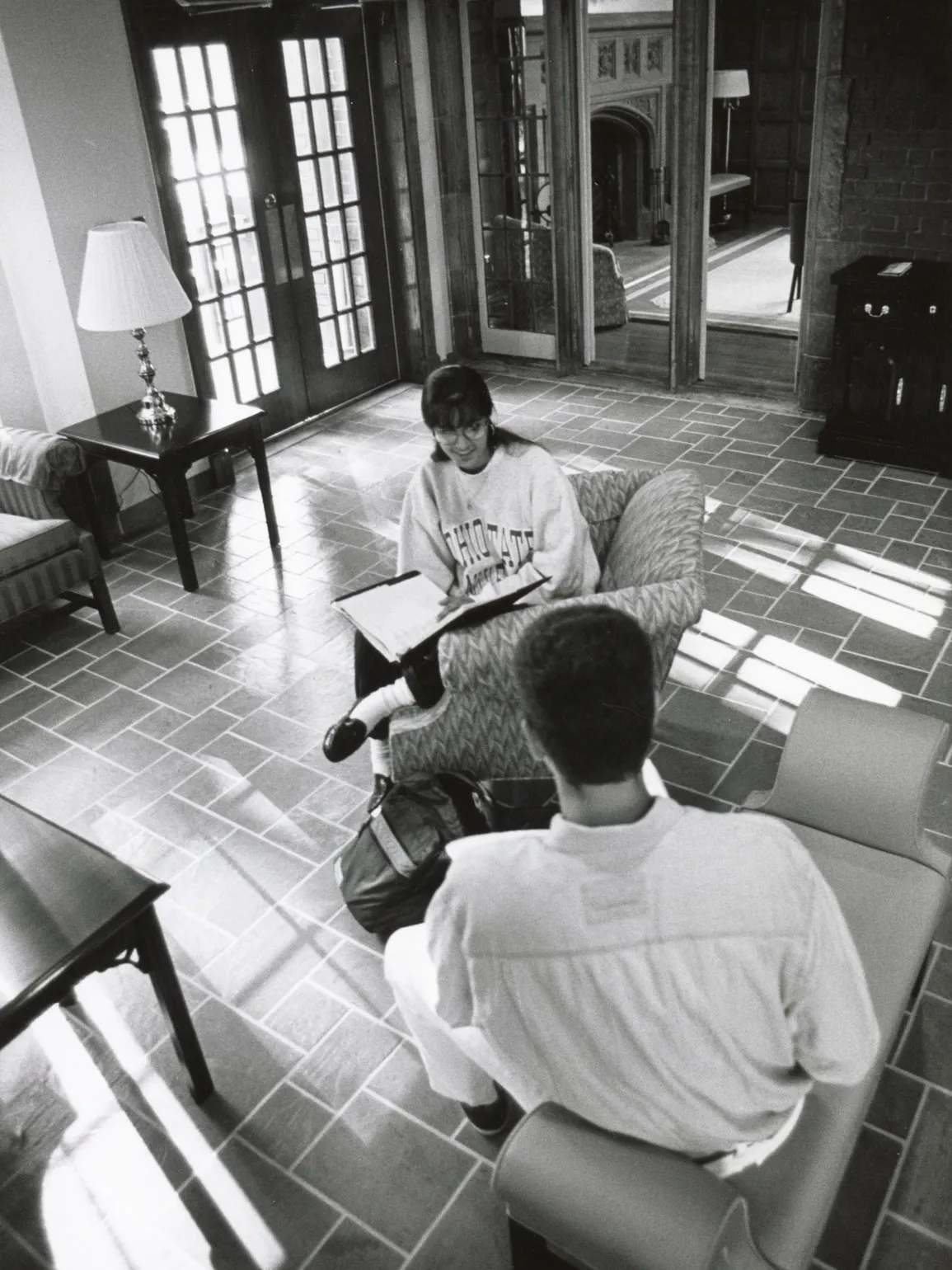 Two people sit in a bright lounge area with tiled flooring and large windows. One person sits in an upholstered chair with a notebook open on their lap, while the other sits on a low chair facing them with a bag at their feet. The room includes a table with a lamp, glass doors leading outside, and reflections in interior windows showing an adjacent room with wood-paneled walls and furniture.