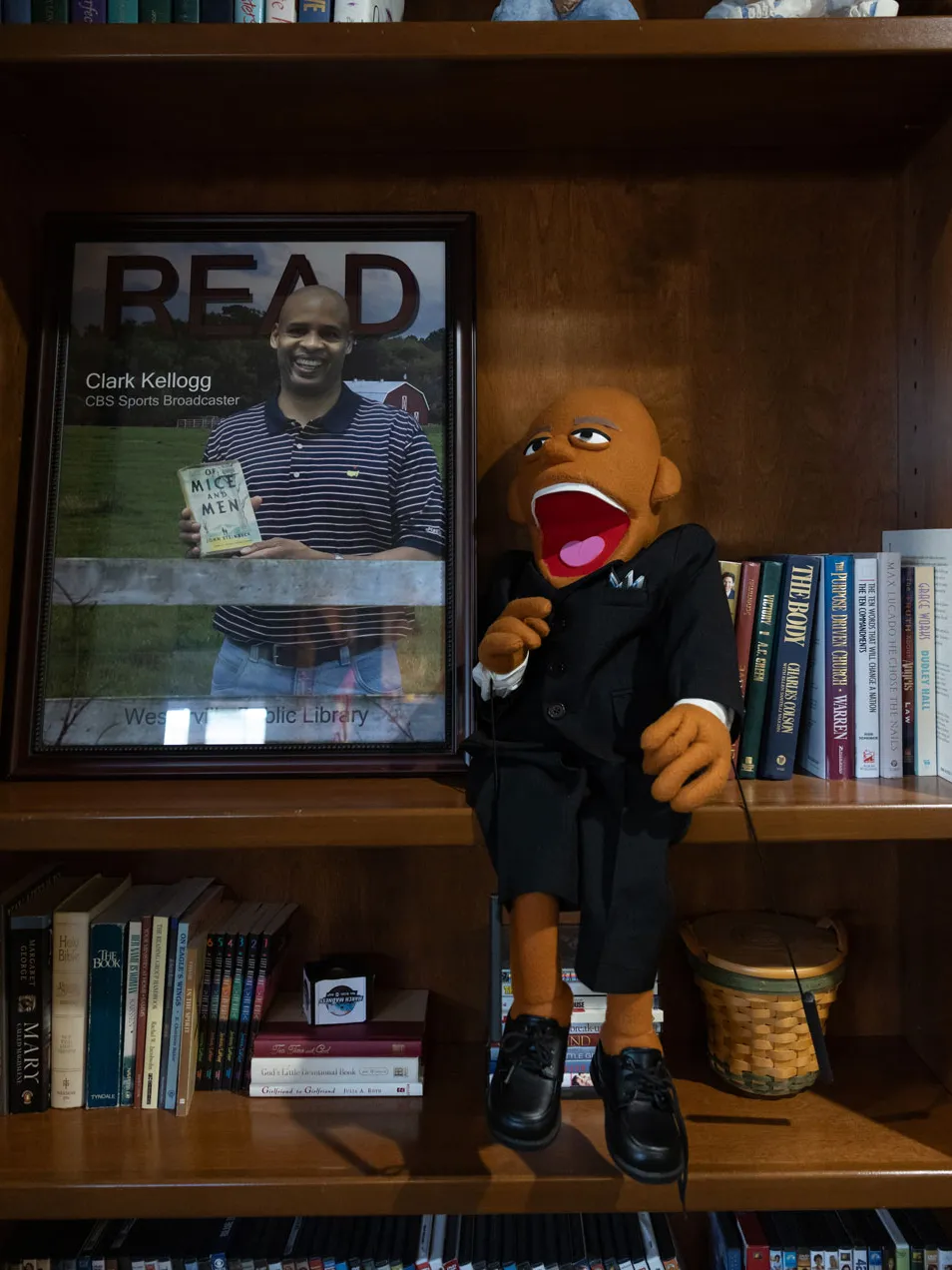 On a shelf at his home, a puppet of Kellogg wears a suit, a big grin and sits on a wooden bookshelf surrounded by books. Next to it is a framed ‘READ’ poster featuring Kellogg holding a book.