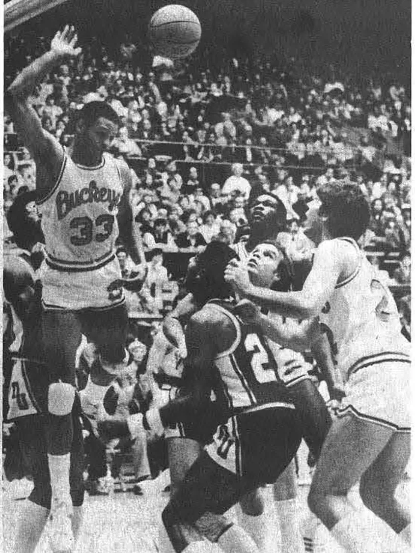 Wearing a Buckeyes jersey, Kellogg is coming back down after leaping near the basket with one arm extended upward. Several opposing players crowd beneath the basket, looking up toward the play, while another Buckeyes player stands close to the action. The scene takes place on a packed court with spectators in the background.