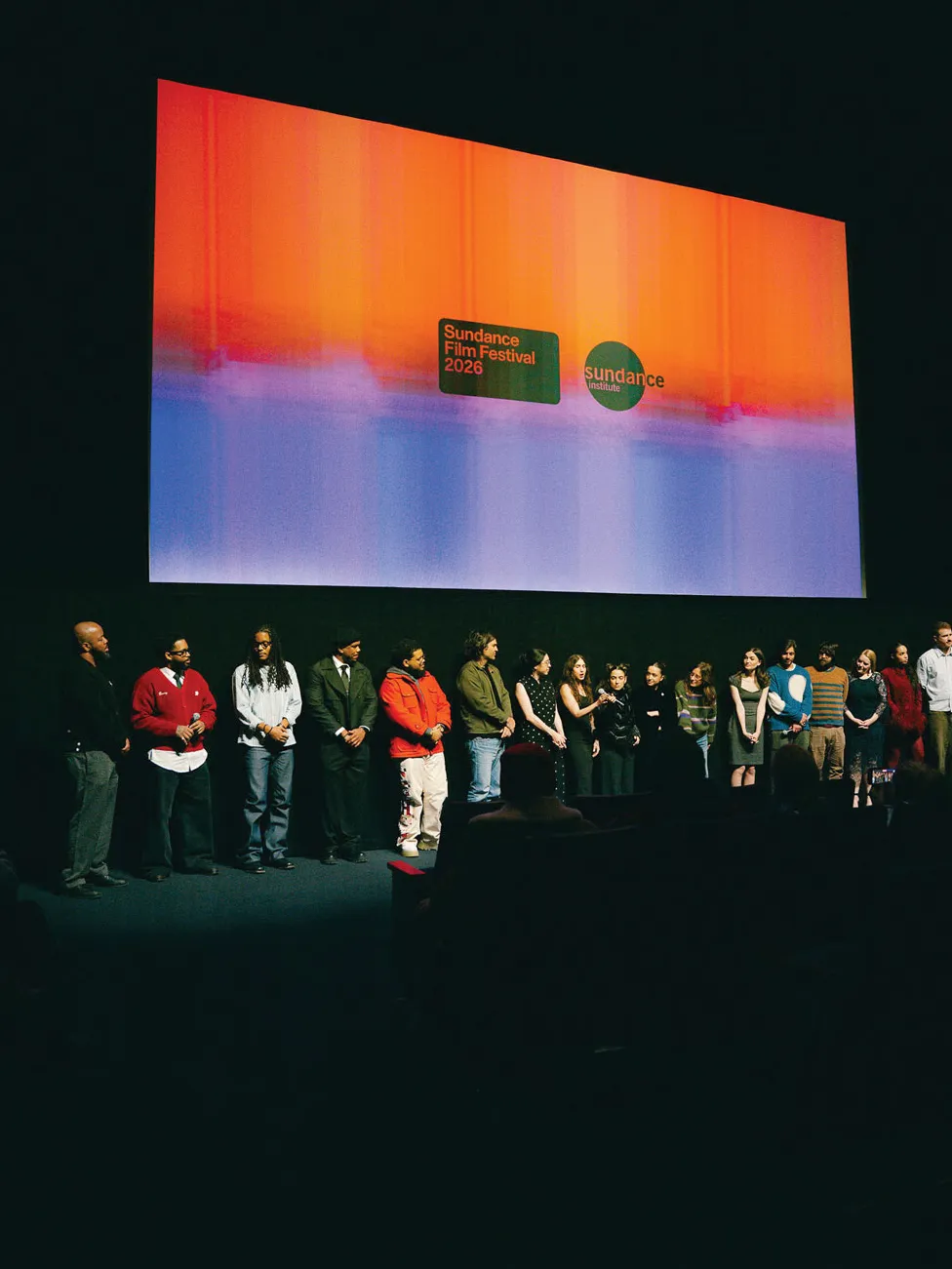 In a dark theatre, the cast and crew of Freelance line up below a movie screen for the Sundance screening. It has two Sundance Film Fetsival logos.