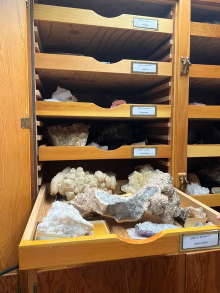 A wooden cabinet has pull-out drawers filled with various minerals