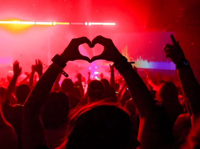 Person making a heart with their hands in a crowd of people dancing by a stage with red lights and fog