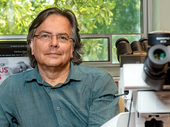 Photo of white man with glasses and blue button up shirt next to a microscope