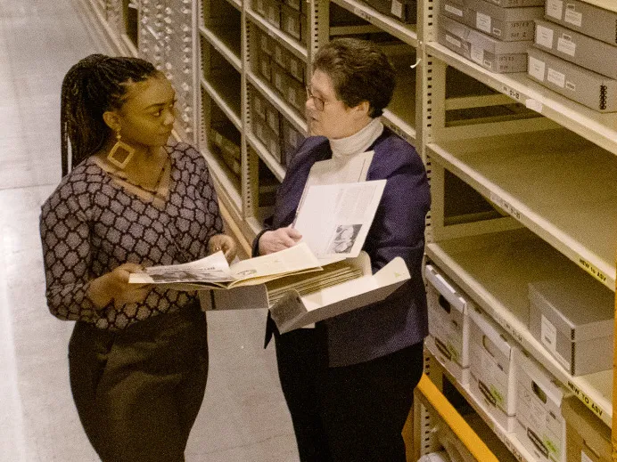 Two women are having a discussion in an aisle between tall metal shelves in a warehouse
