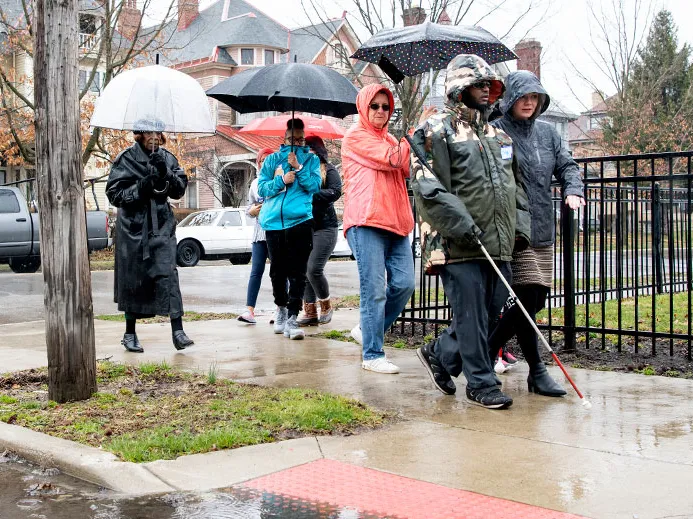 group of people walking in the rain behind a blind man using a cane
