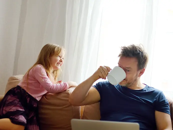Father drinking coffee while working on the computer and his kids laughing and playing beside him