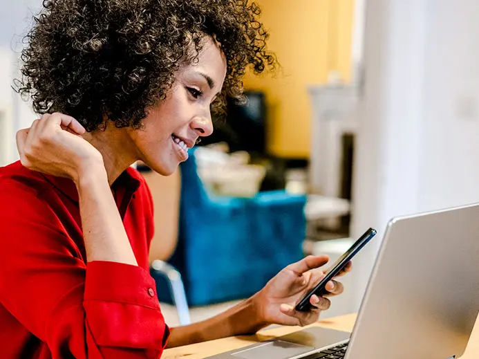 woman with curly hair wearing a red shirt is holding her phone while working on the computer