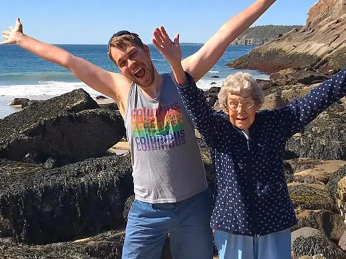 Brad and his Grandma Joy are standing next to one another on a rocky beach with their arms outstretched