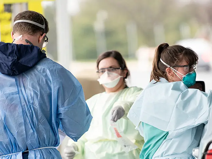 A man and two woman, all wearing hospital gowns, face masks, and gloves, assist in drive-up COVID-19 testing