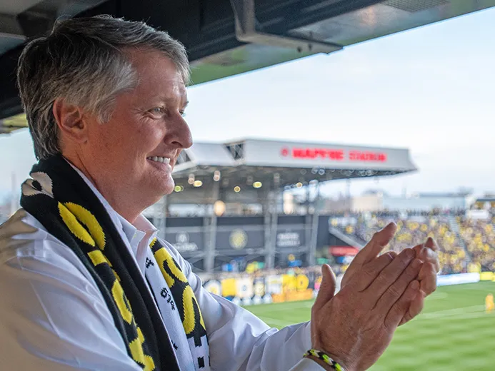 Dr. Pete Edwards is clapping for Columbus Crew SC from his seat inside Mapfre Stadium. He is wearing a black and yellow scarf and a white long sleeve shirt.