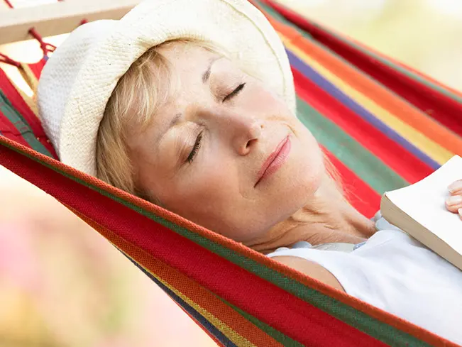 woman wearing a hat and a sun dress with a book folded on her chest while taking a nap on a colorful hammock