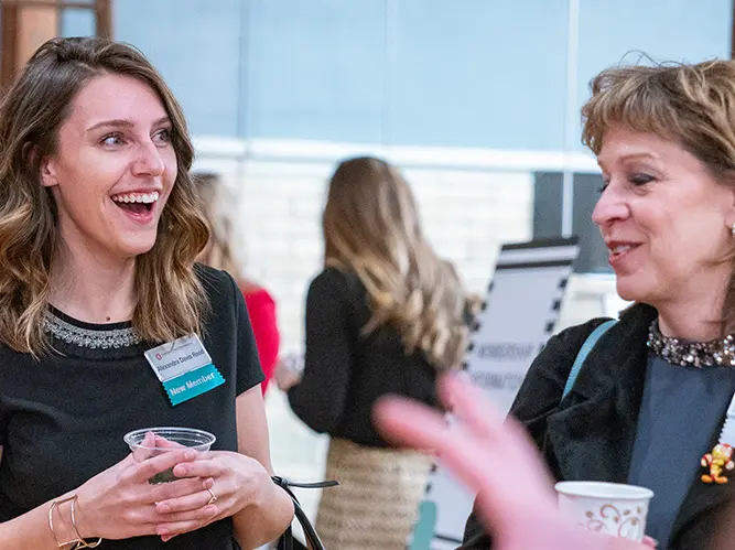 Two women are in a discussion while at an event. They are wearing names tags and holding disposable cups., More than 200 women comprise the membership of Women & Philanthropy, a giving circle-style philanthropic organization dedicated to funding researc