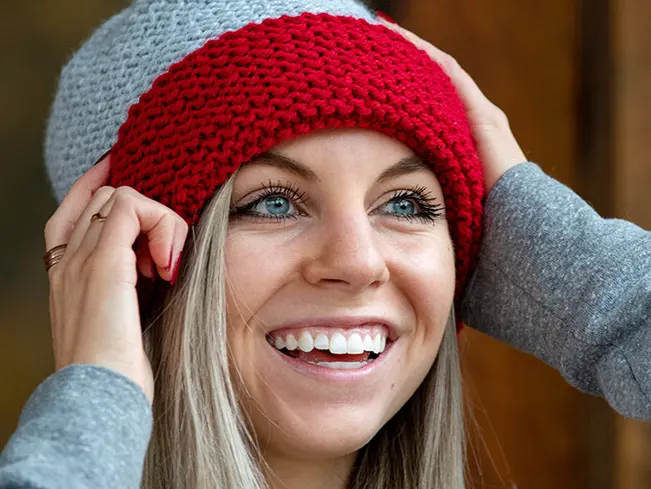 A young woman is wearing a gray and red knit winter hat and smiling