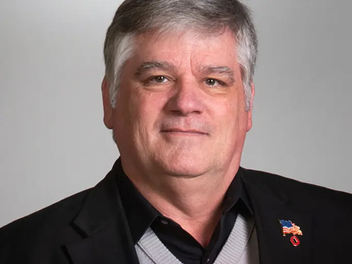 Portrait of Kelley Crooks wearing a dark suit jacket and a united states flag pin on his lapel