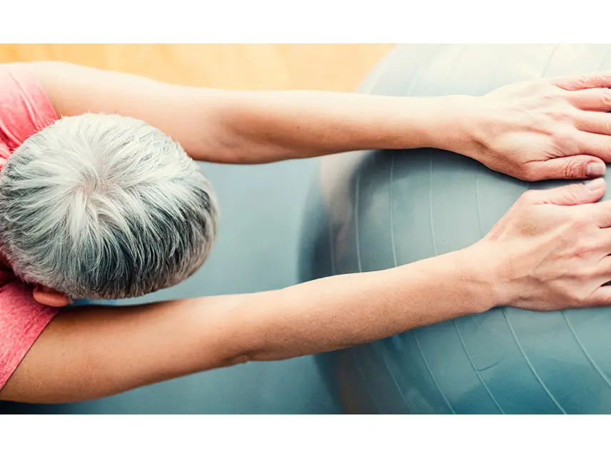 A woman with short gray hair is kneeling on a yoga mat with her hands resting on a teal exercise ball and her arms straight