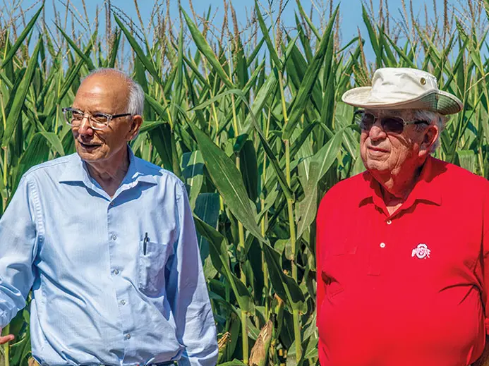 Two men are standing in front of corn stalks. One is wearing a light blue long sleeve button-up and the other is wearing a red polo shirt and a tan hat.