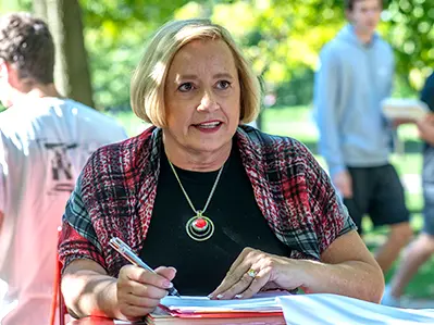 A mature woman seems invested in listening to a person off-screen. She is outside and taking notes on a packet on top of a stack of folders and notebooks.