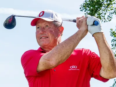 An older white man in a scarlet golf shirt finishes the arc of his golf-club swing behind his head. He&#039;s looking into the distance, with a slight smile, as if he&#039;s proud of where the ball landed.