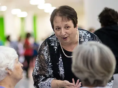 A woman smiles as she leans forward to engage with a pair of older adults. They are shown from behind while she faces the camera.