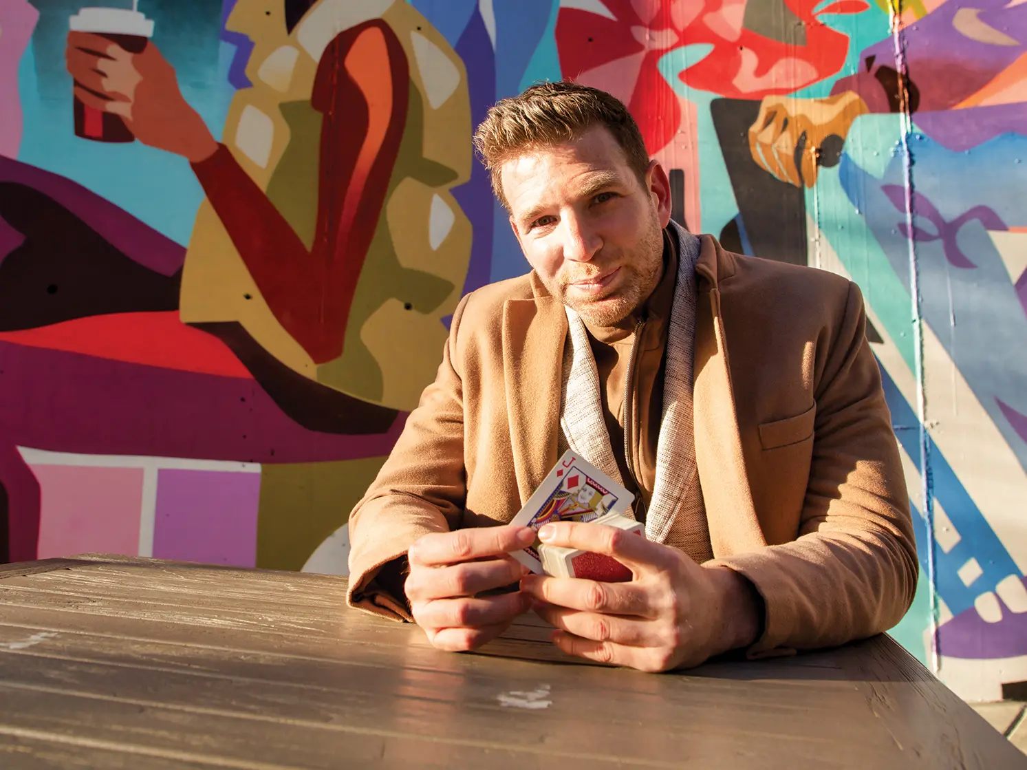 Smiling with his eyes, Joshua Jay holds a jack of diamonds while sitting at an outside mural