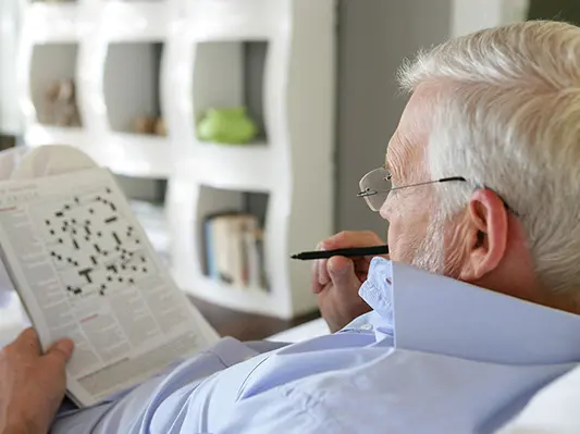 An older man is seated in a chair while doing a crossword puzzle with a pen.