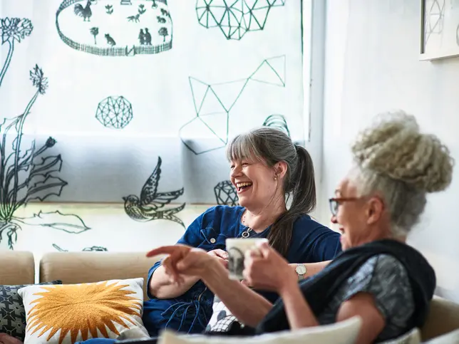 Two woman sit at home laughing, enjoying coffee, and each others company
