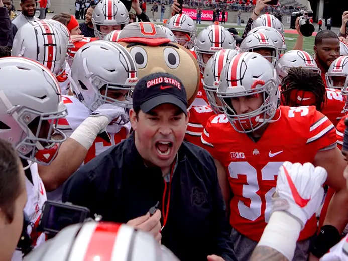 Head coach of the Ohio State football team is standing in the middle of a huddle of players in football uniforms. He is wearing a black long sleeve shirt and a black OSU hat.