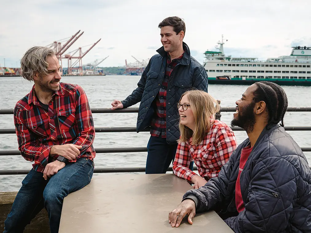 Four people, three wearing red plaid, laugh together around a table on the waterfront. A big ferry and cranes can be seen behind them.