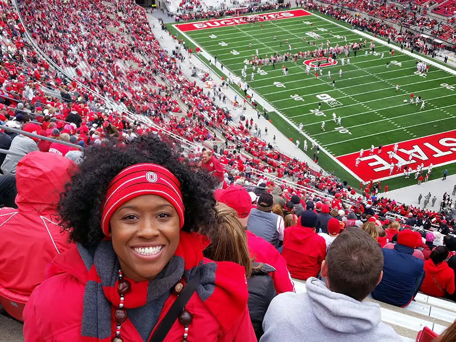 Inside the ’Shoe, a happy woman decked out in Ohio State gear sits high in the stands as football players line the field behind her.