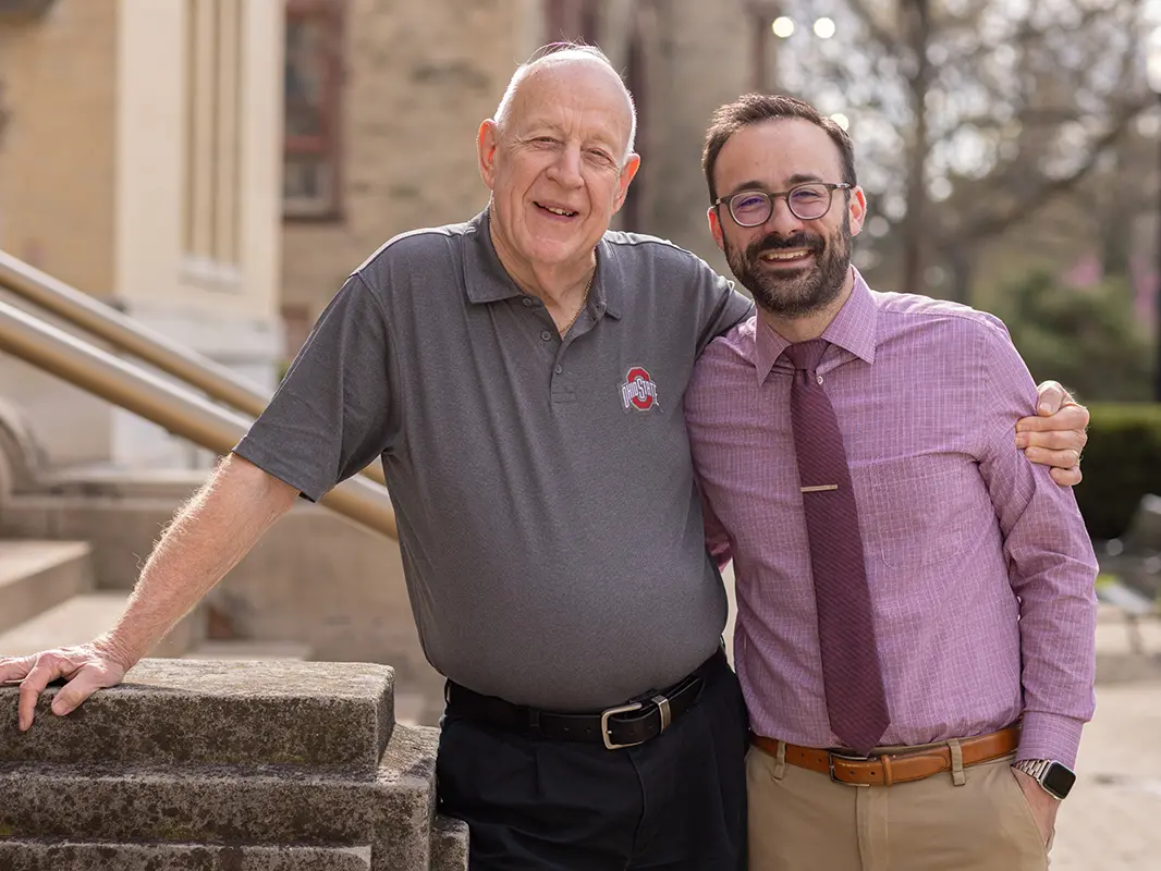 An older man in an Ohio State golf shirt and a younger man wearing a button-up shirt and tie stand side by side with arms around each other. They look relaxed and happy.