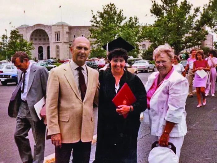 In an old color photograph, a young woman in cap and gown stands between her proud parents. Ohio Stadium can be seen in the background.