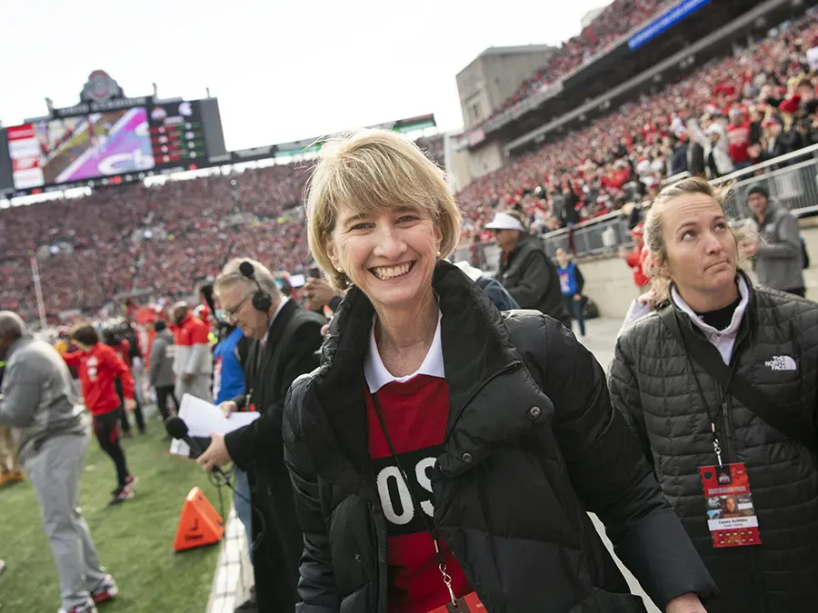 Ohio State President Kristina M. Johnson, a white woman with short blond hair, smiles cheerfully from the sidelines of a fall football game.