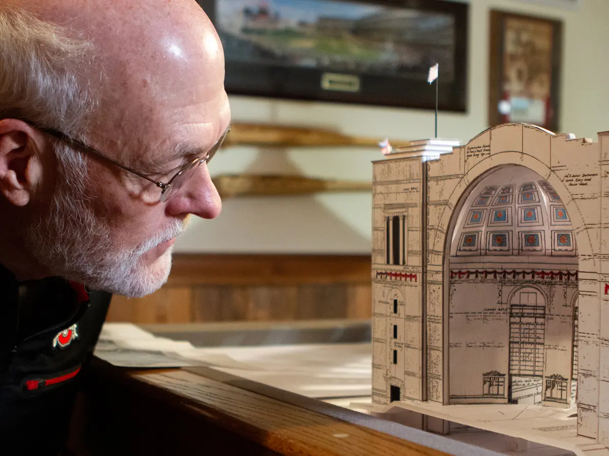 In a basement decked out with Ohio State keepsakes, an older man in glasses takes a close look at the front of the Ohio Stadium model he built, which is slightly taller than his head.