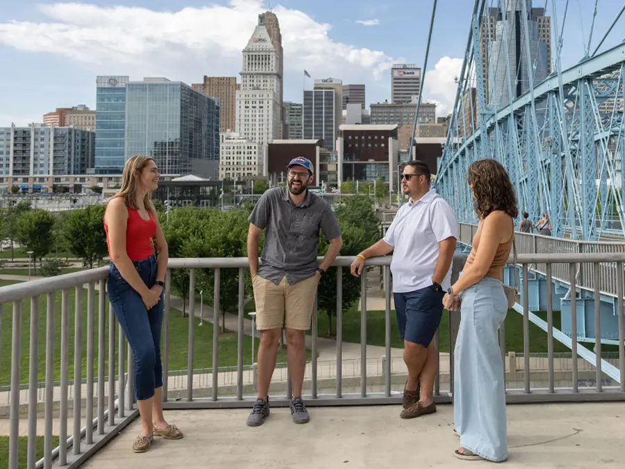 Four Buckeye alumni in their 30s, laughing and wearing summer clothes, make eye contact as they talk. In the distance are a blue-green bridge, part of the Cincinnati skyline and fluffy white clouds.