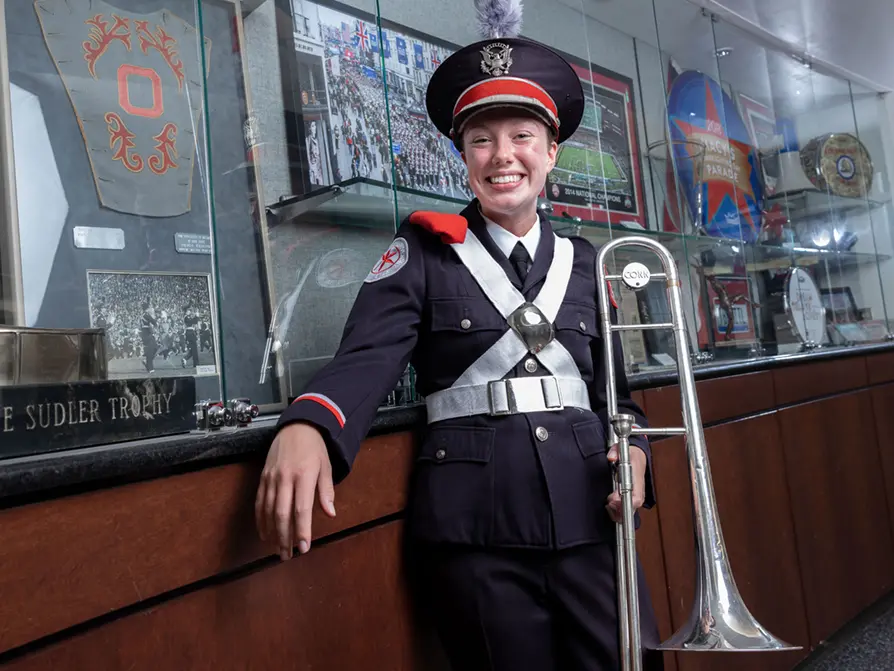 A woman in a marching band uniform smiles brightly as she leans on a counter in front of display cases of TBDBITL memorabilia.