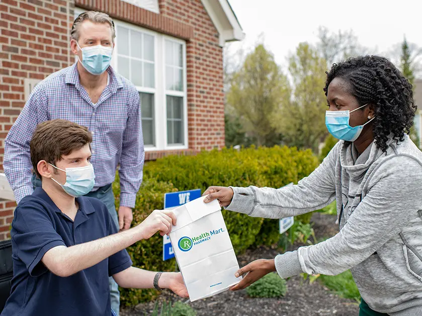 A lady with hands a pharmacy bag to a young man and his father. All wear surgical masks.
