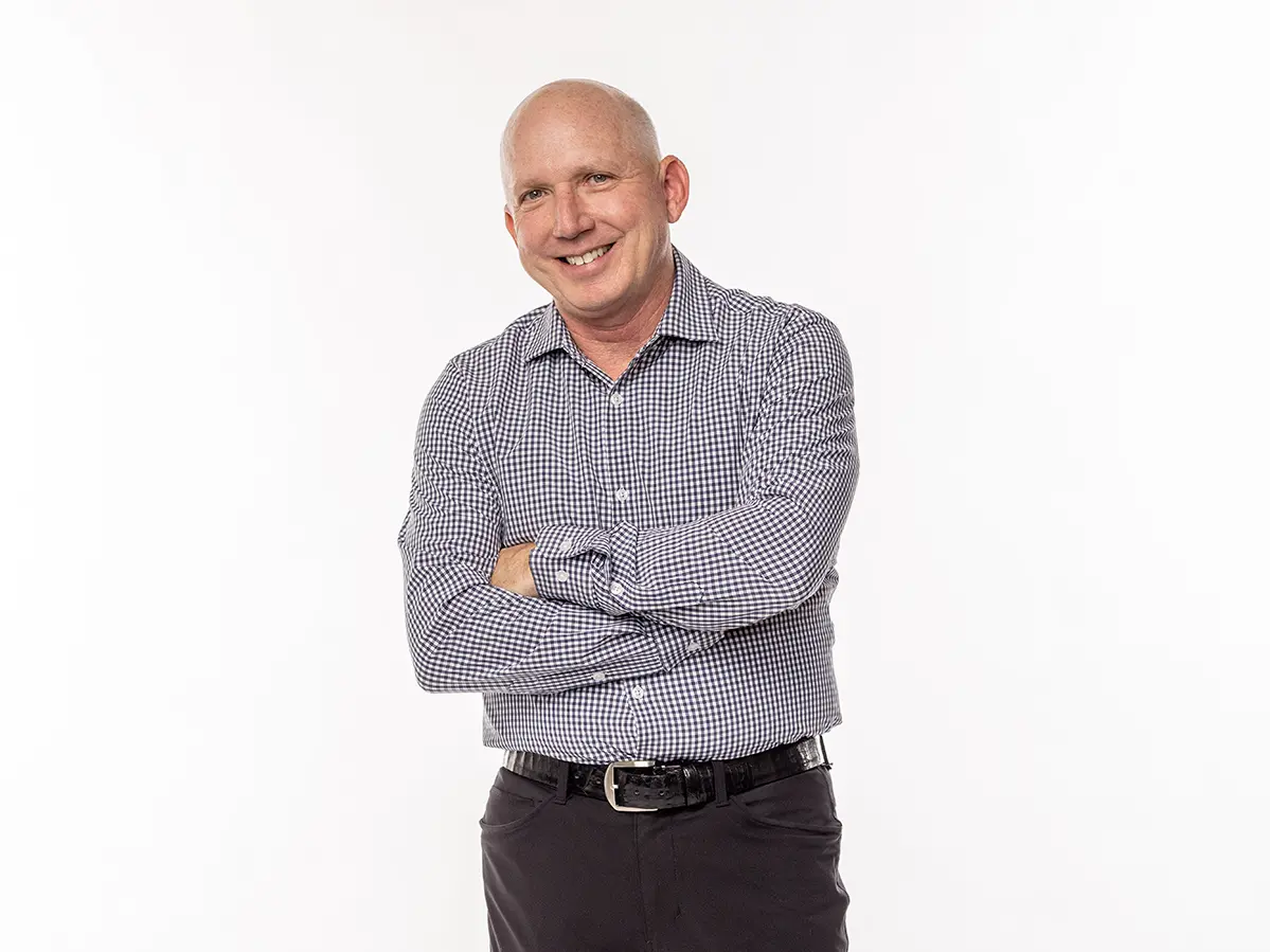 A white man with a shaved head stands confidently with a big smile and arms crossed.