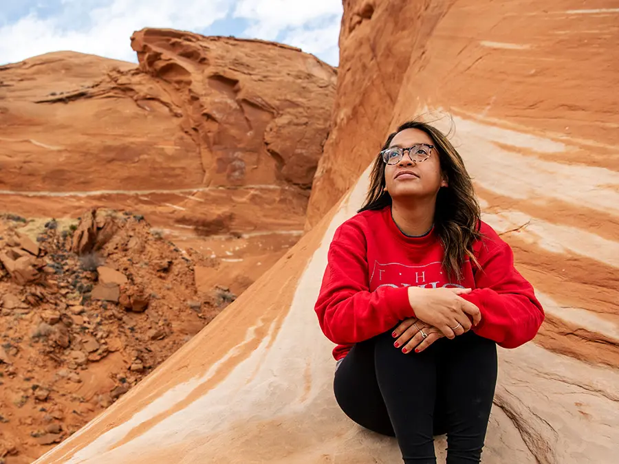 A woman in a red Ohio State sweatshirt looks to the sky as she sits on a giant, moon-shaped, orange and cream-colored rock formation. She’s significantly higher than the ground, but behind her, portions of the rock go much higher, off the top of the photo.