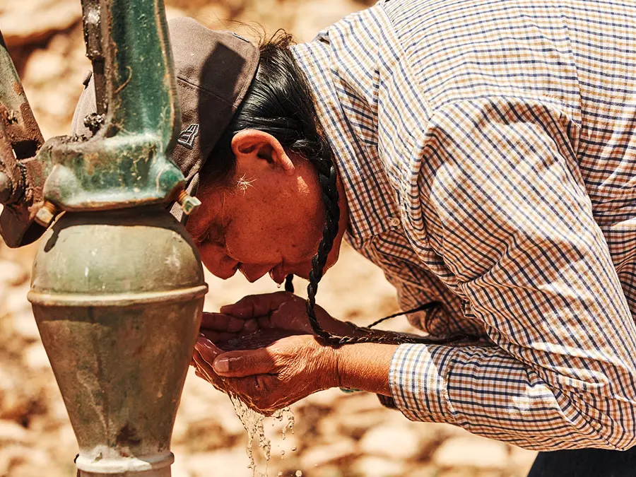 A Navajo Nation man wearing braids and a ballcap bends over to drink water from his own cupped hands, which he just pumped from a green metal well apparatus covered by a thin layer of dirt.