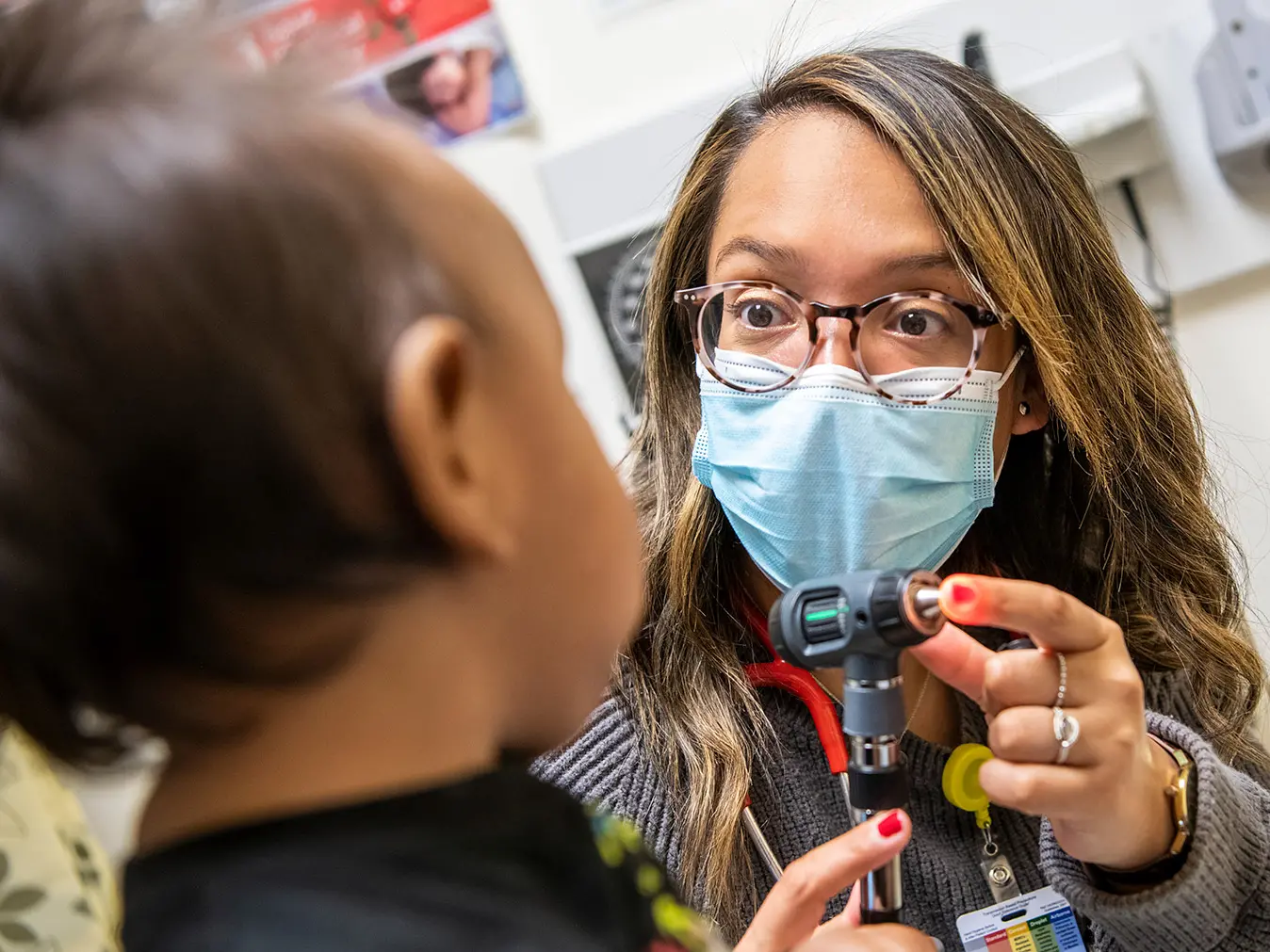 A young doctor of Guatemalan descent wears round tortoiseshell glasses, a surgical mask and a wide-eyed expression as she connects with a toddler. She’s showing the little girl the tool she’ll use to look in her ears.