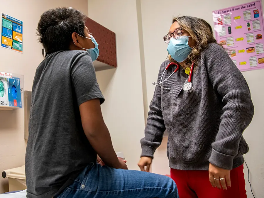 A young doctor in a surgical mask prompts a child patient to raise his shoulders during a medical exam