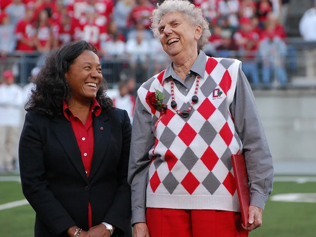On the field in Ohio Stadium, an older white woman wearing a scarlet and gray argyle vest stands next to a younger black woman. She’s looking at her friend and they are both laughing.