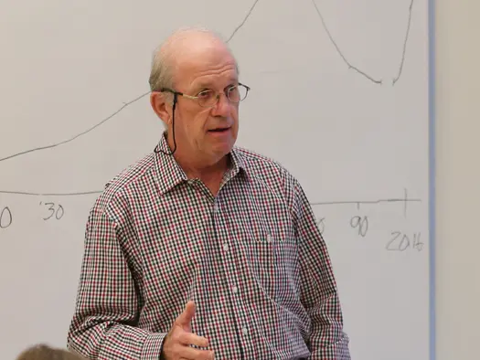 An older man wearing glasses and a checked button-down gestures while teaching a class in front of a white board.