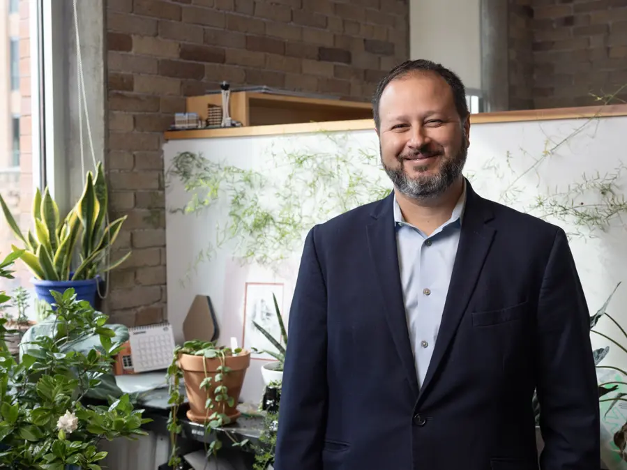 A bearded man in a navy blazer stands smiles broadly in his office, surrounded by plants