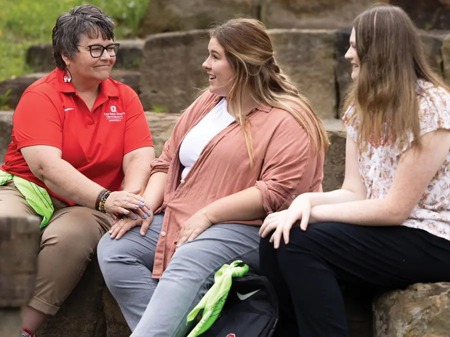 A woman wearing an Ohio State shirt and two students smile at each other in a supportive way as they chat and sit on large stone steps on Ohio State’s Mansfield campus.